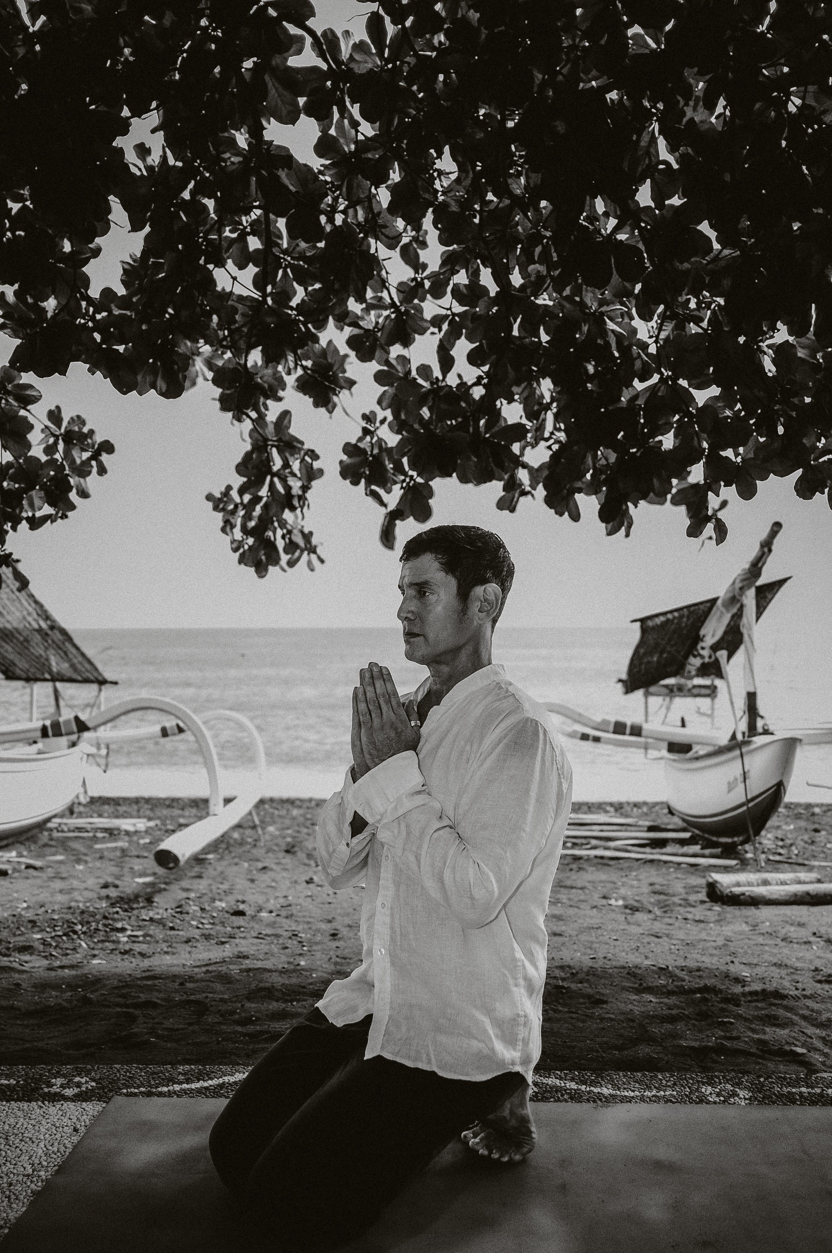 A person kneels on a mat under a tree near a beach, with wooden boats in the background, engaged in a peaceful meditation or prayer pose while wearing a white shirt.