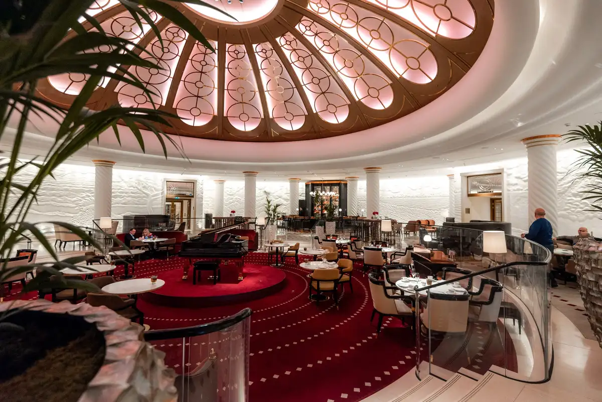 A circular hotel lobby features a large domed ceiling with ornate gold and pink patterns above red carpets and seating.