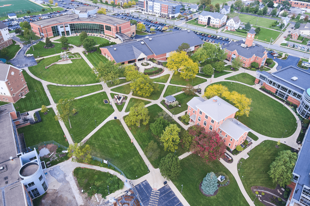 Aerial view of a scenic college campus with lush green spaces, academic buildings, and walkways under a clear sky