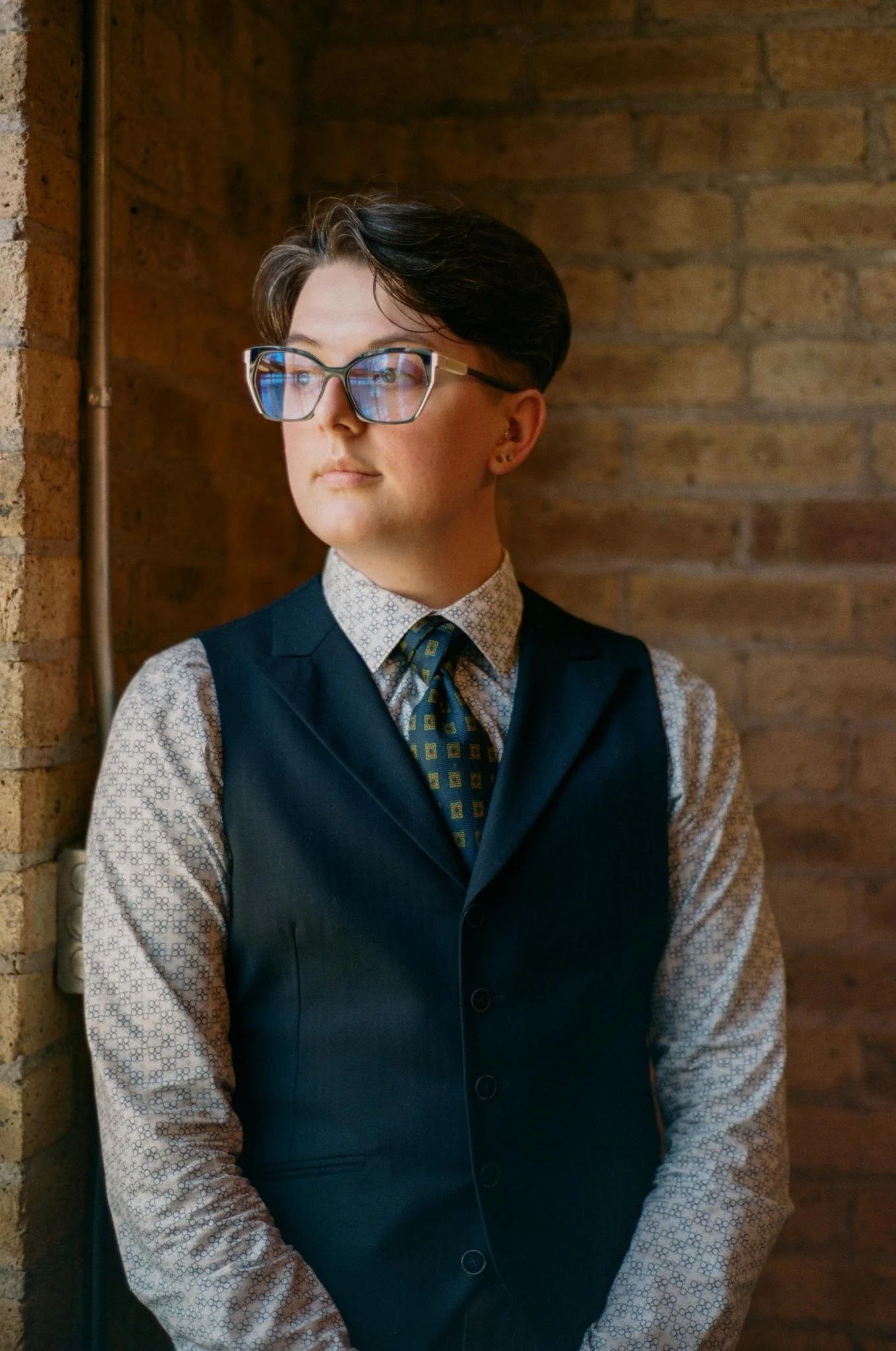 Uma pessoa de cabelo curto e óculos, vestindo uma camisa estampada, gravata e colete escuro, está em pose pensativa diante de uma parede de tijolos aparentes, olhando para o lado.