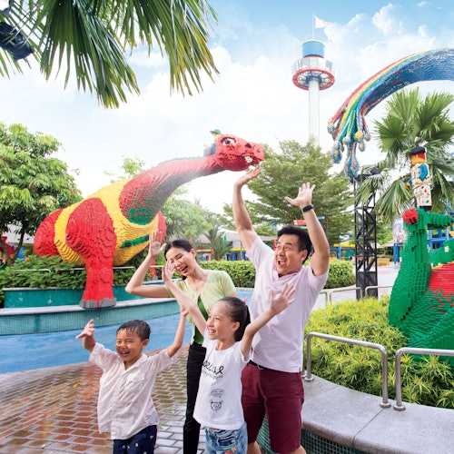 A smiling family of four joyfully raises their hands near colorful Lego sculptures, including a red and green dinosaur, in a park setting.