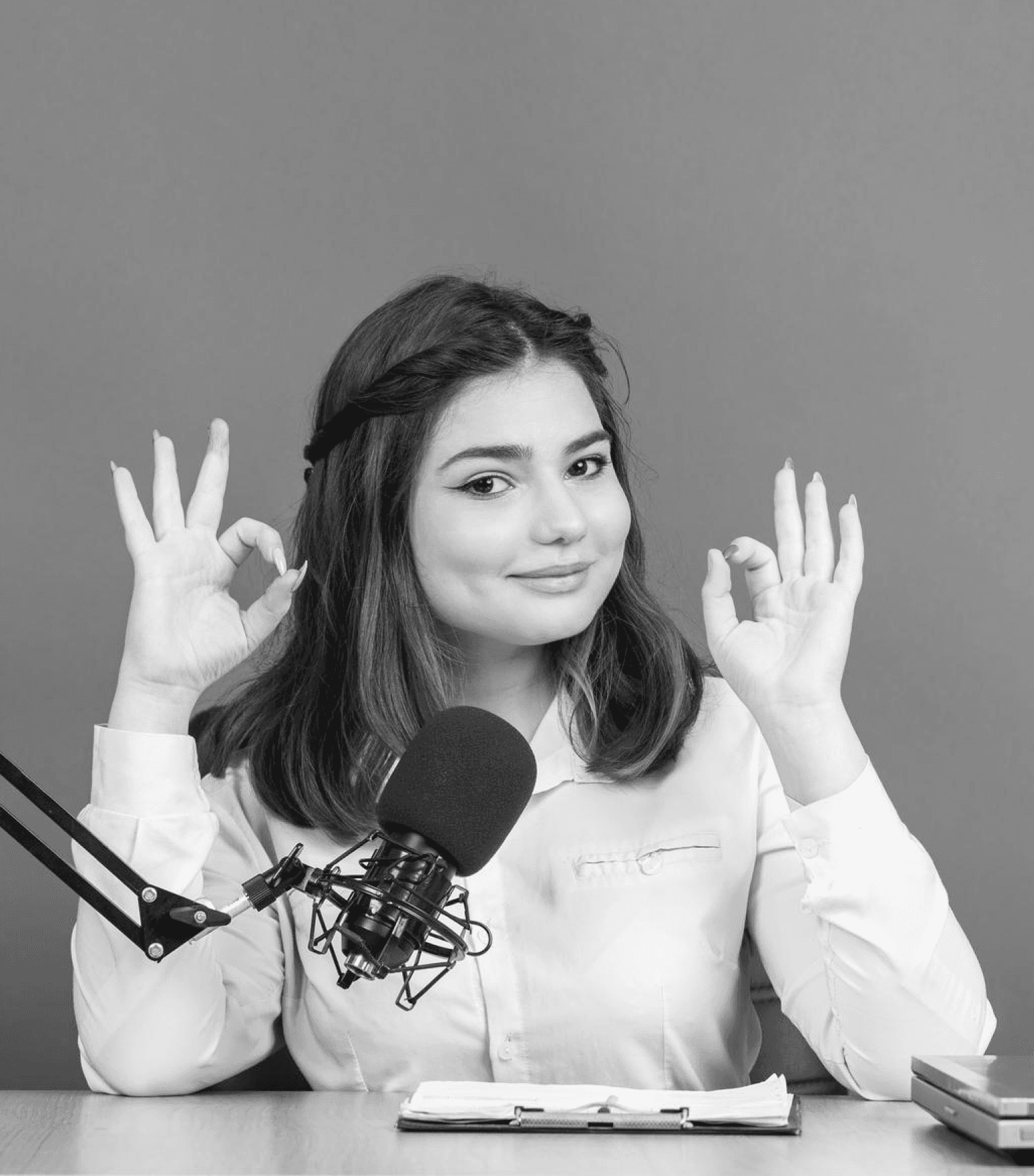 a young woman smiling, sitting in front of a podcast microphone, and making 'OK' signs with both hands.