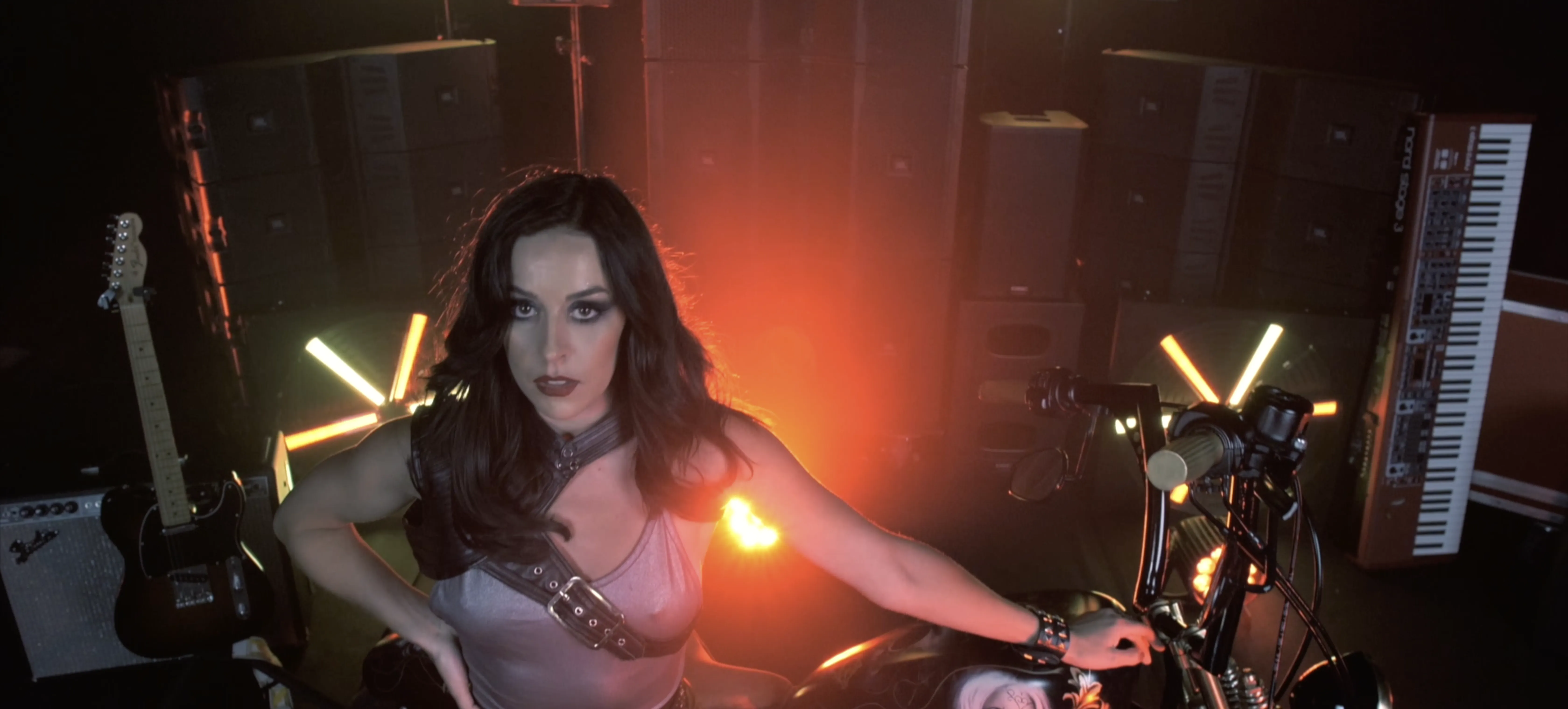 A woman with long dark hair, wearing a light-colored outfit, poses confidently on a motorcycle amidst a backdrop of illuminated stage lights and musical instruments, including guitars and keyboards, creating a dramatic and energetic atmosphere.