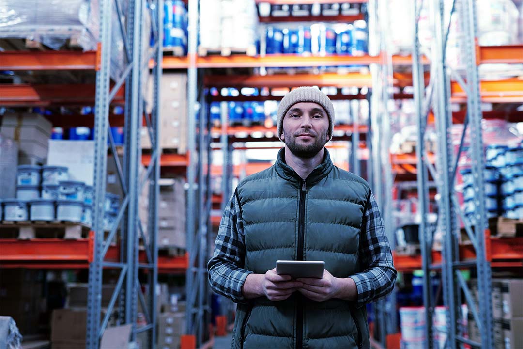 A man standing in a warehouse holding an electronic tablet, looking directly at the camera with a confident expression. The background features shelves stacked with boxes, suggesting an industrial or logistics setting.