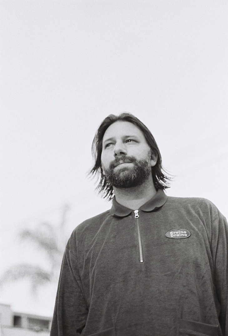 Black and white photo of Dominic Maker, acclaimed producer and member of Mount Kimbie, known for collaborations with artists like James Blake, Travis Scott, and Jay-Z. Maker stands outdoors, looking into the distance, wearing a zip-up shirt with a 'Braino Draino' logo.