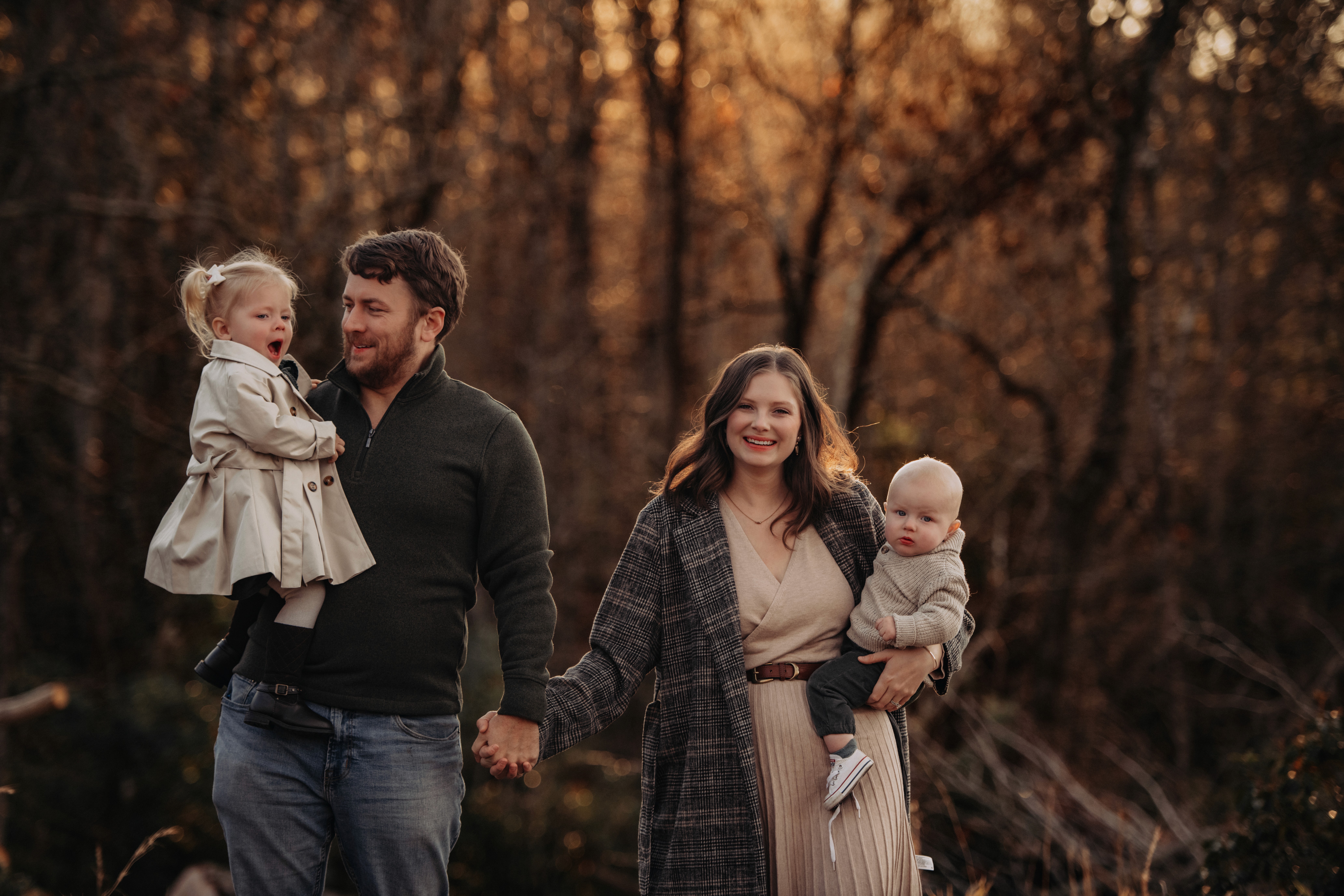 A family of four smiles as they walk through a park holding hands. A woman and man hold children, all looking happy.