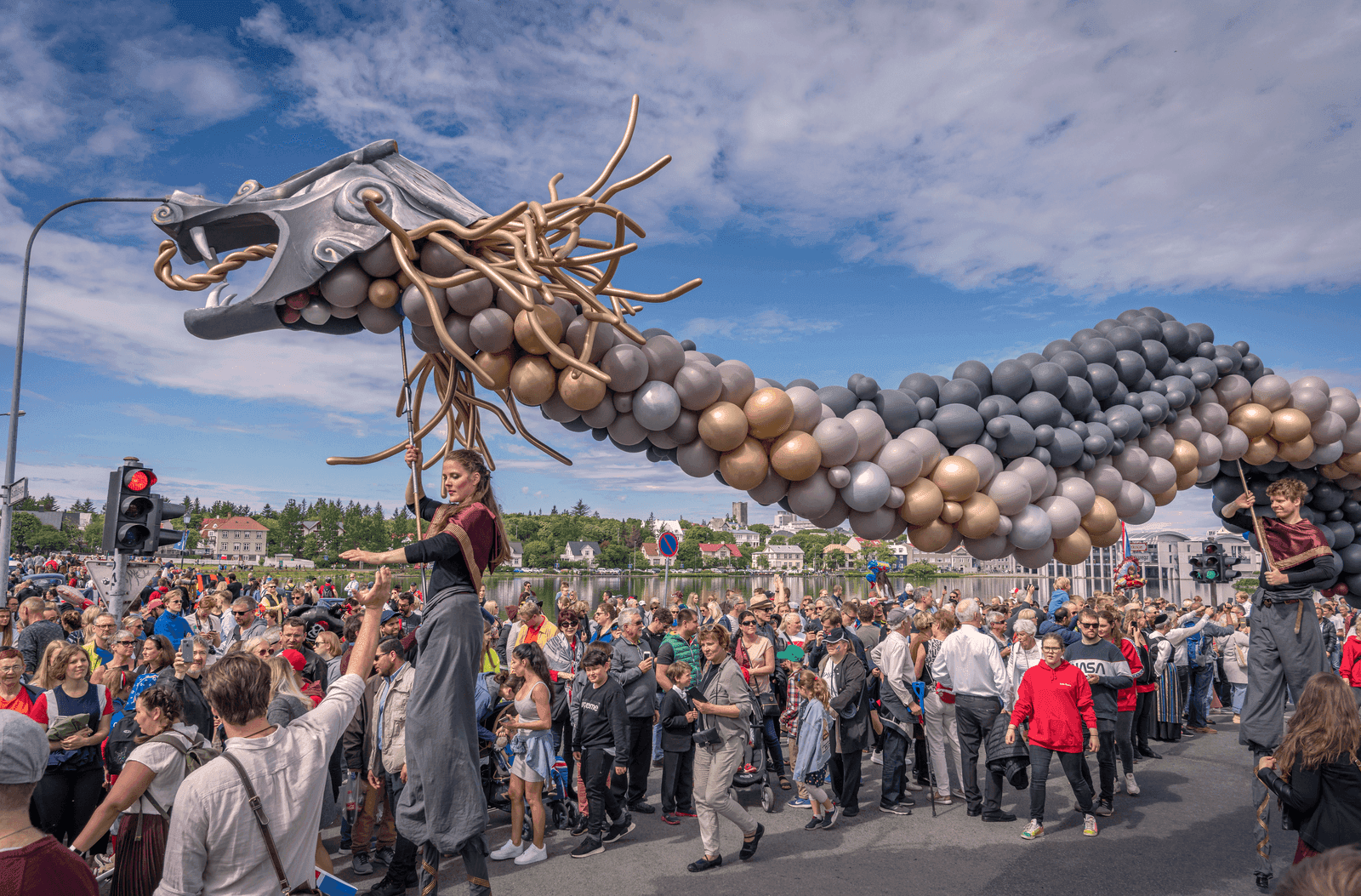 An outdoor scene from Iceland's National Day on June 17 in Reykjavík. A diverse crowd of spectators watches a street parade next to the city lake. Two performers wearing dark, stylized costumes walk on tall stilts, holding up a long, winding dragon parade prop constructed from metallic gold, silver, and charcoal grey balloons. The sky is blue with light clouds.