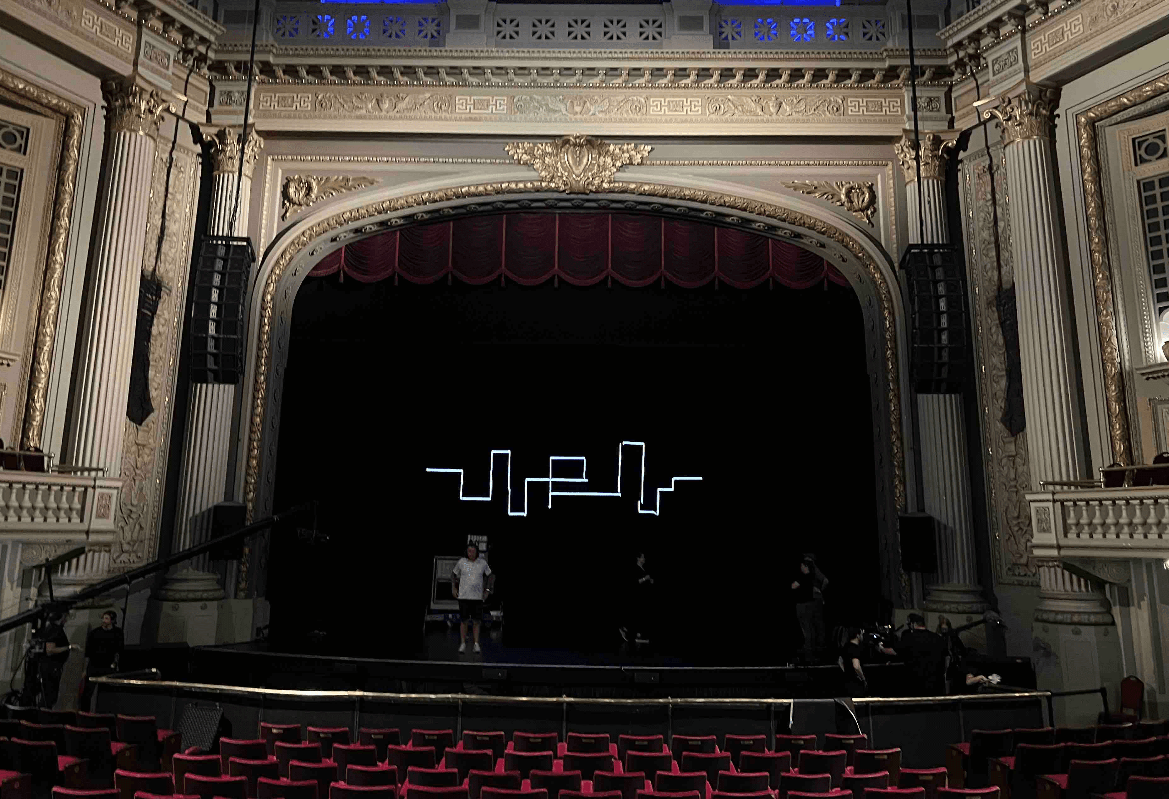 Interior of a theater featuring ornate architecture, a dark stage, and a blue ceiling illuminated with lights.