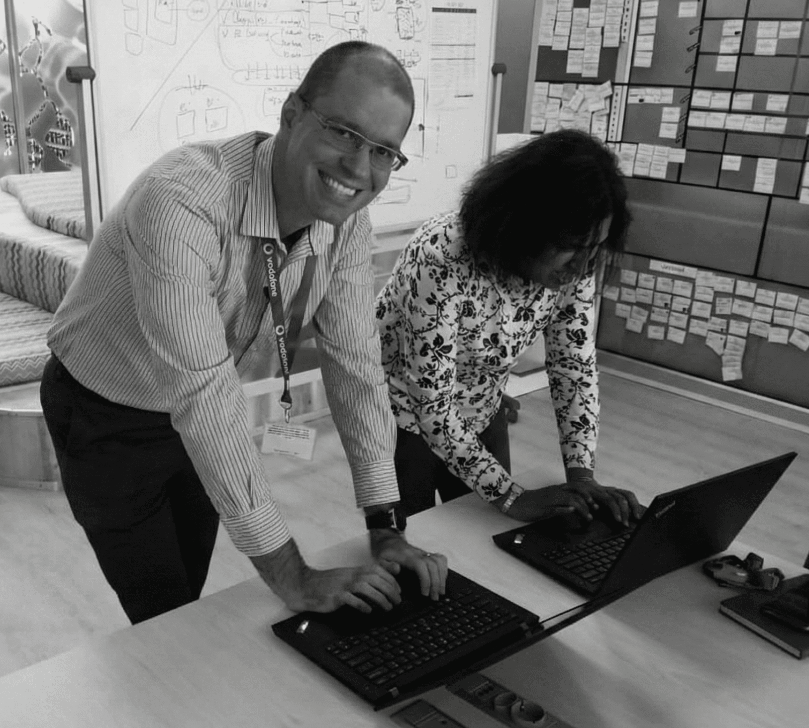 A young person with curly hair smiles at the camera while sitting in front of a laptop against a dark background.