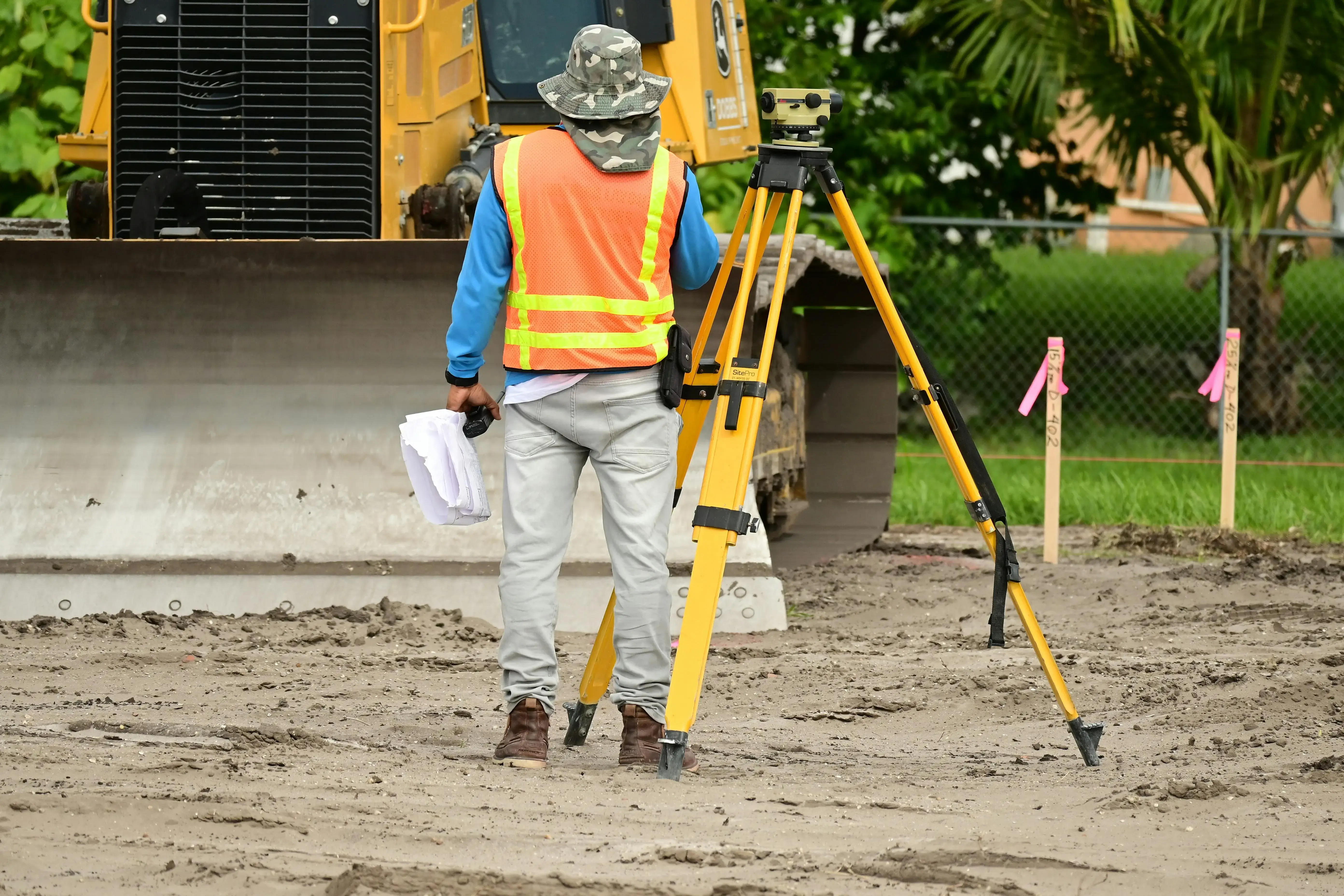 Construction worker in a neon vest and hat stands next to a yellow tripod on a muddy site, facing a bulldozer. Pink flags and greenery in the background.