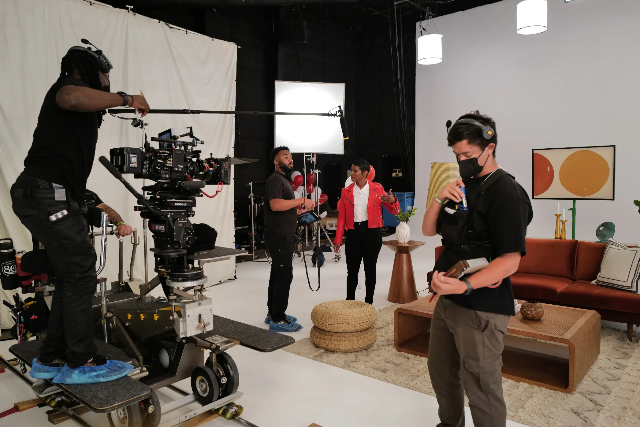 A film crew works on a set with cameras, lights, and sound equipment. One person holds a boom mic, another wears headphones, and a woman stands near a sofa with modern decor. Crew members wear masks and casual clothing.