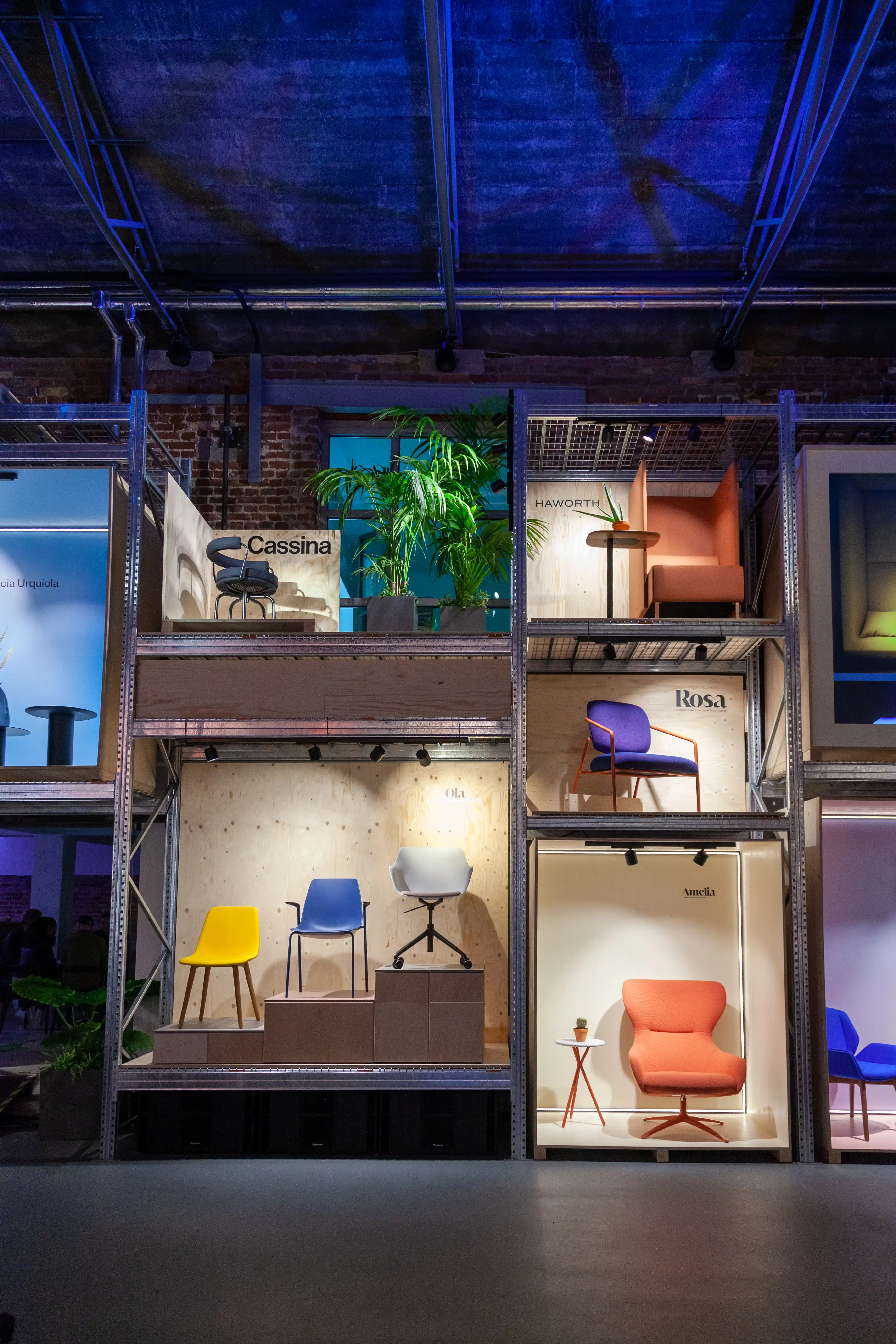 Multi-level steel shelving system at the Haworth ORGATEC booth presenting colorful chairs and armchairs in lit display boxes.