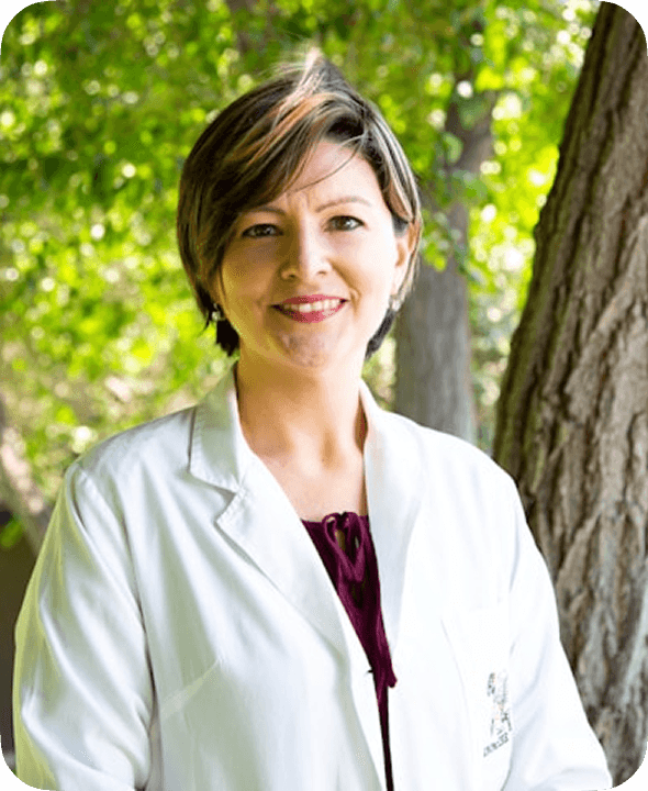 A smiling person wearing a white lab coat stands outdoors against a backdrop of lush green trees, conveying a sense of professional expertise and connection to nature.