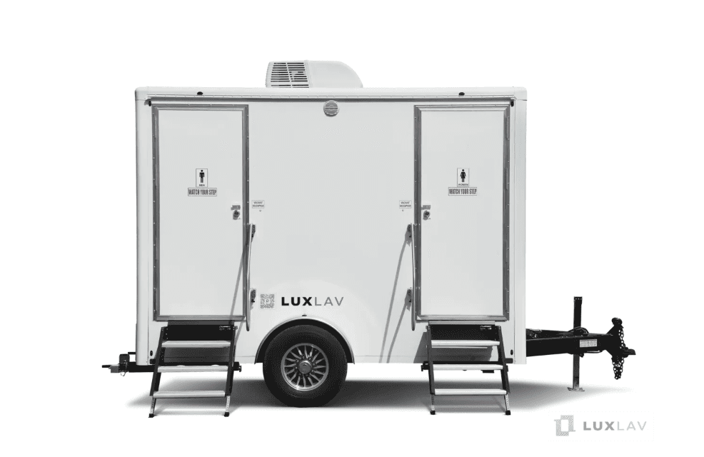 White two-door restroom trailer with wooden entry steps on grass at dusk, a city skyline rising behind it