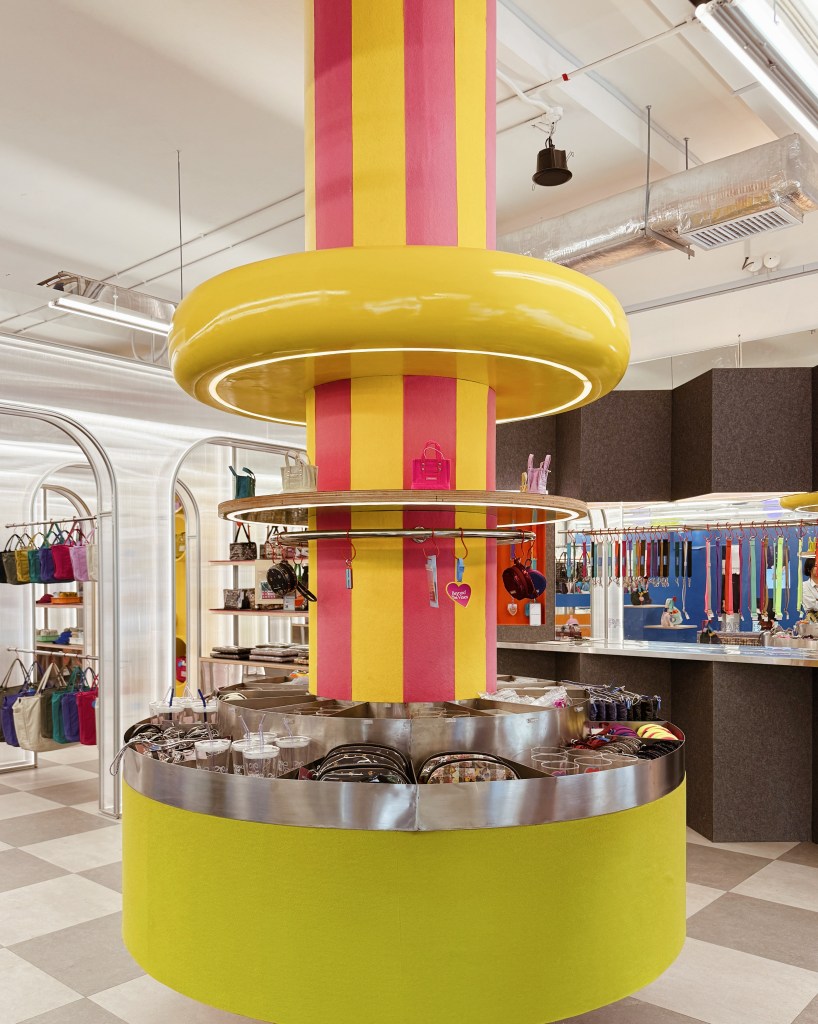 A colorful retail display featuring a vibrant yellow and pink cylindrical stand at the center, surrounded by various bags and accessories in a trendy shop.