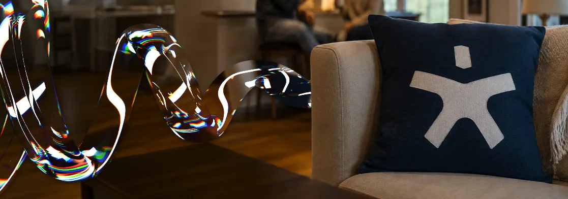 Fotografia de um ambiente interno com duas pessoas conversando desfocadas ao fundo. Em primeiro plano, à direita, um sofá bege acomoda uma almofada azul escura com um logotipo da spark maxx em branco. À esquerda, flutuando no ar, há uma forma abstrata em 3D parecida com uma fita de vidro retorcida e transparente, refletindo feixes de luz coloridos com um efeito digital.