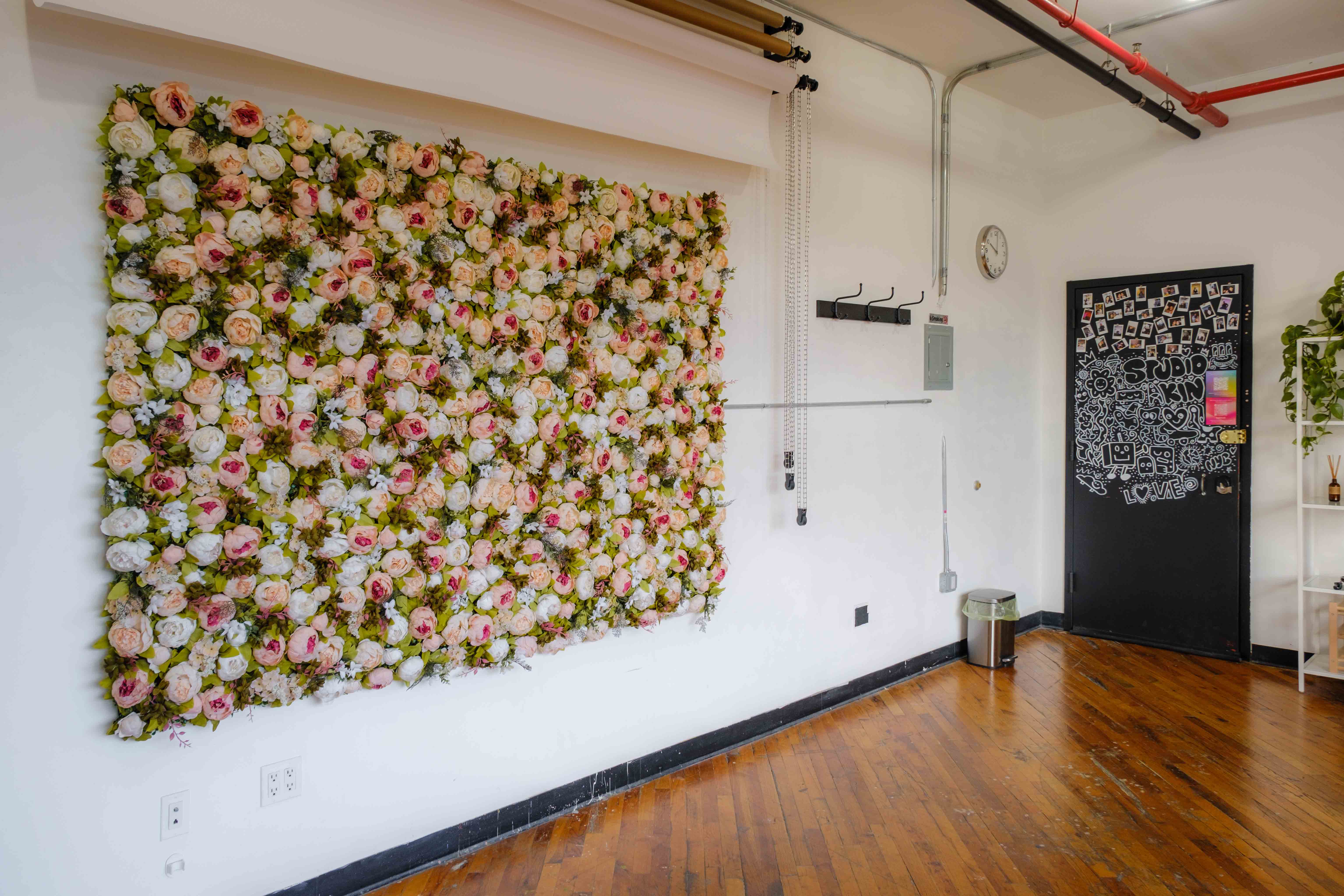 A photo studio wall adorned with a large, vibrant floral installation, creating a striking backdrop. The room also features hardwood floors, a chalkboard-style door decorated with doodles, and a setup for background rolls.