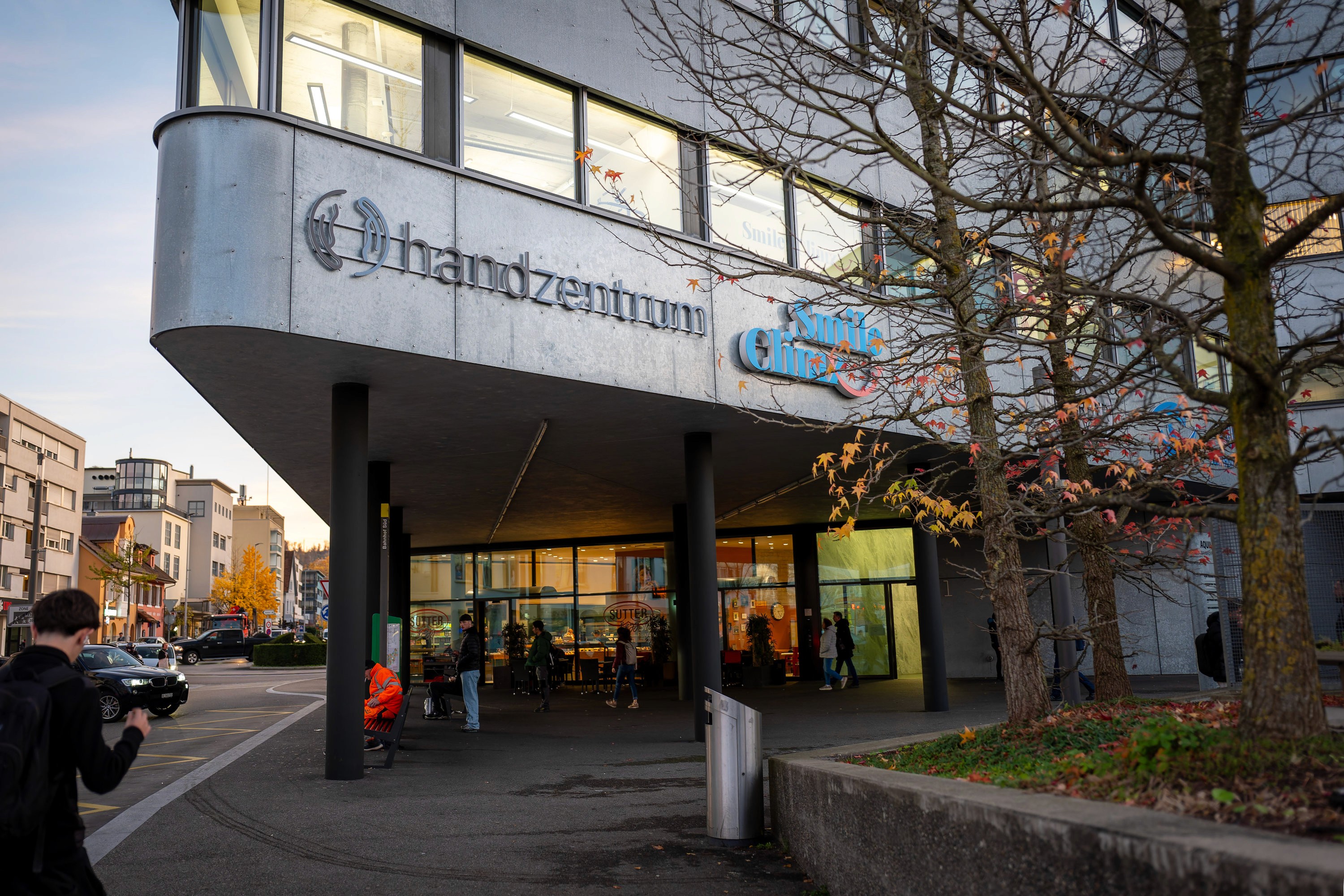 Der Aquila-Tower in Pratteln: Ein modernes Gebäude mit großen Fenstern und dem Logo des Handzentrums über dem Eingang, Menschen, die draußen sitzen und spazieren gehen, und ein Baum mit spärlichem Laub im Vordergrund.