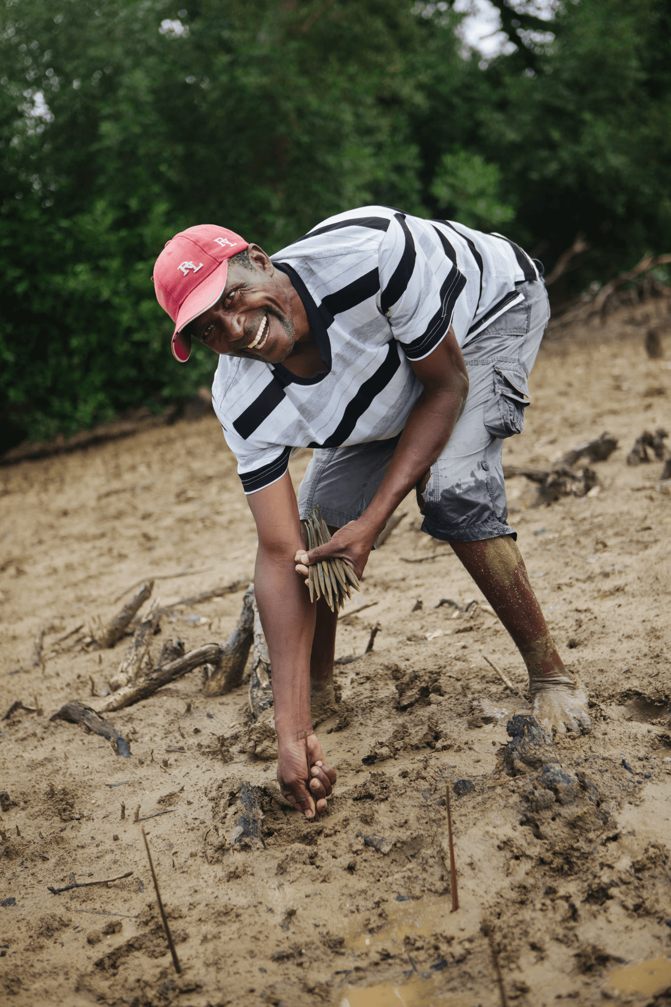 Lachende man met een rode pet en gestreept shirt die jonge bomen plant in modderige grond, wat de betrokkenheid van de lokale gemeenschap in bunq's herbebossingsinspanningen benadrukt.