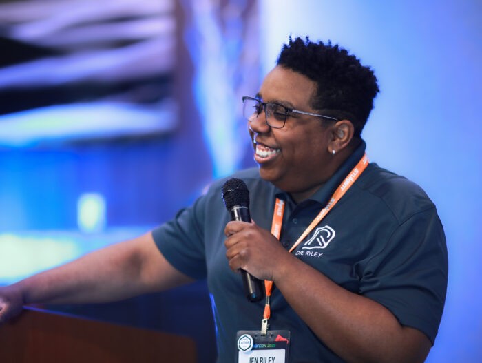 Smiling speaker in dark polo shirt with event logo holds a microphone and gestures during a presentation. They wear glasses and an orange lanyard with a conference badge. Blue-toned background is softly blurred.