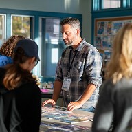 The club chairman is preparing materials for a group activity in a community room, members are interacting in the background.