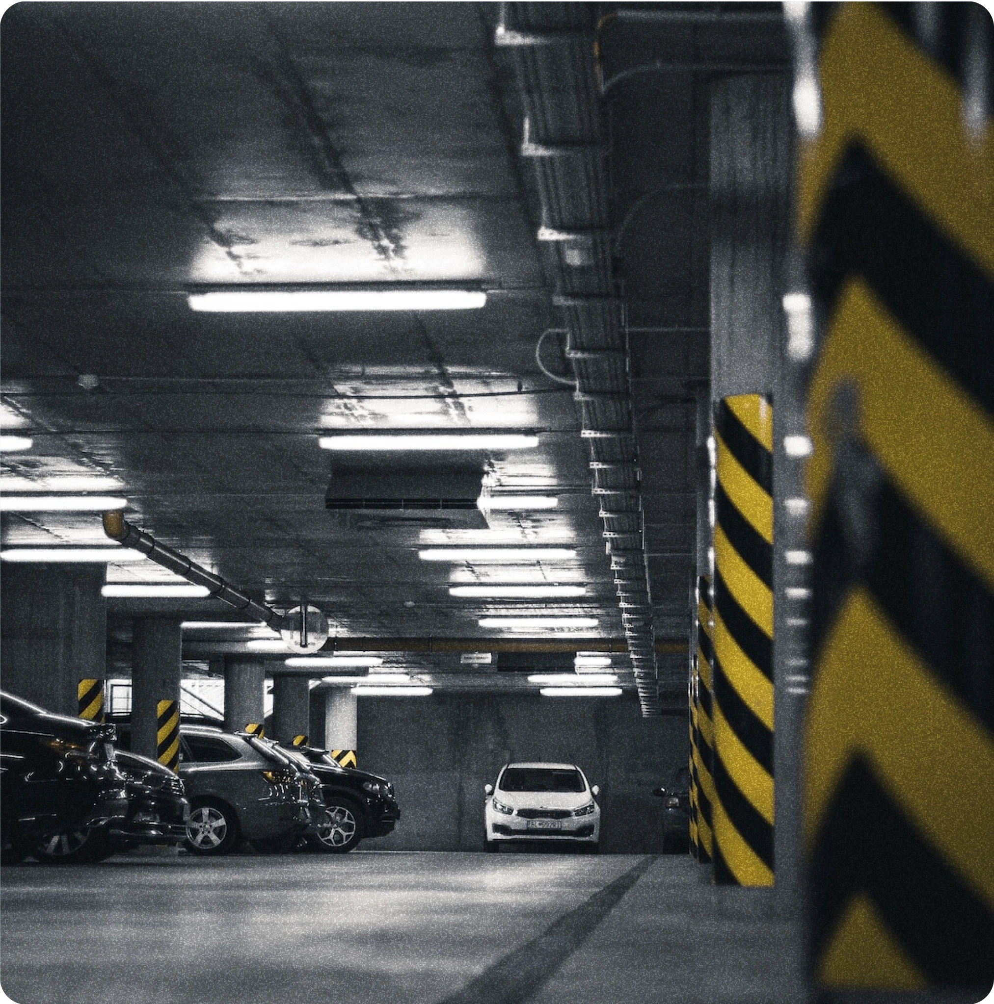 A dimly lit underground parking garage with several parked cars. The ceiling is lined with fluorescent lights, and the concrete pillars are marked with black and yellow hazard stripes. The foreground shows empty parking spaces, while the background reveals a white car parked in the distance.