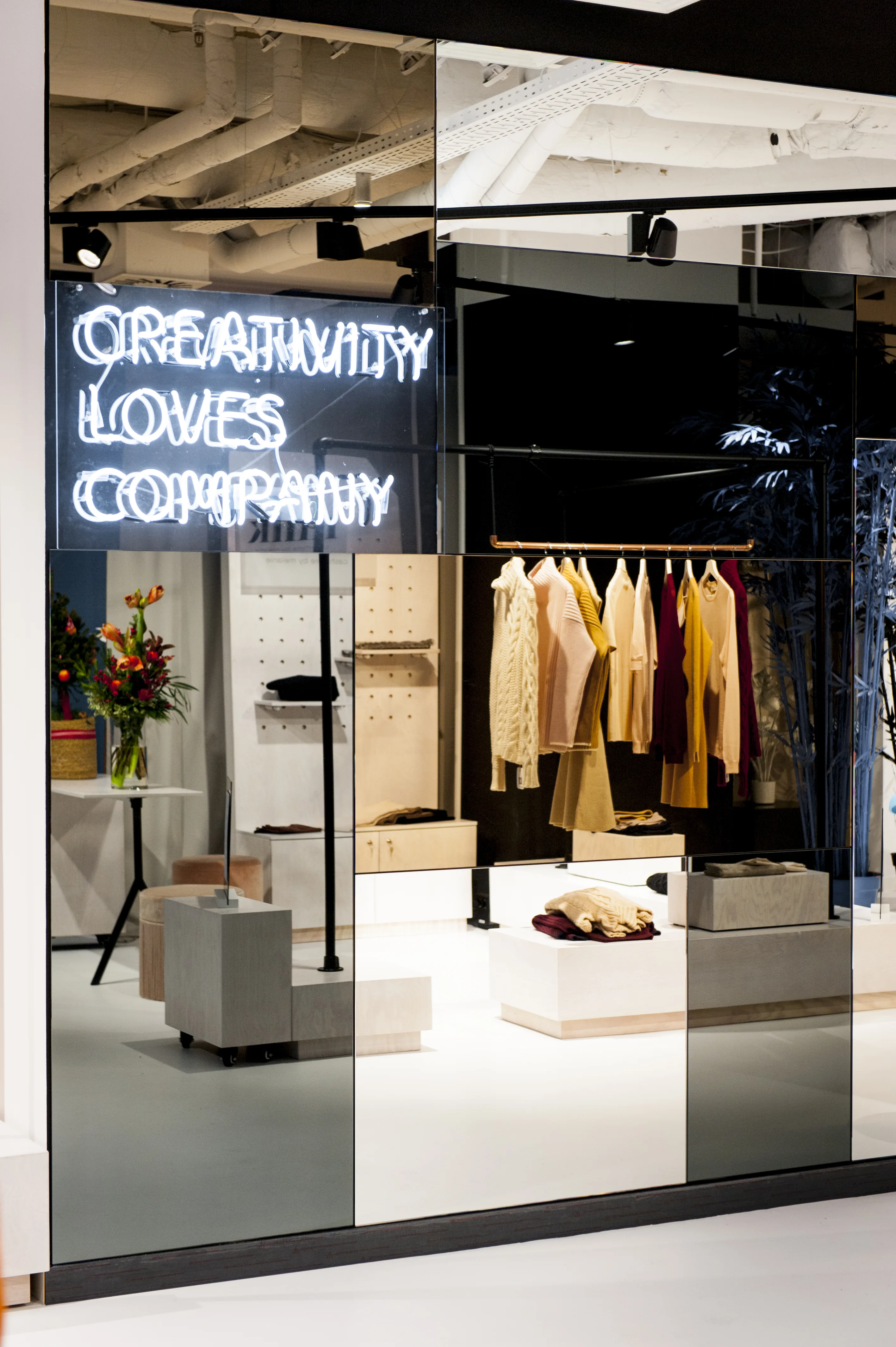 Mirrored box at FREIRAUM SHOP with the white neon slogan Creativity Loves Company above a curated fashion display with hanging knitwear in beige tones.