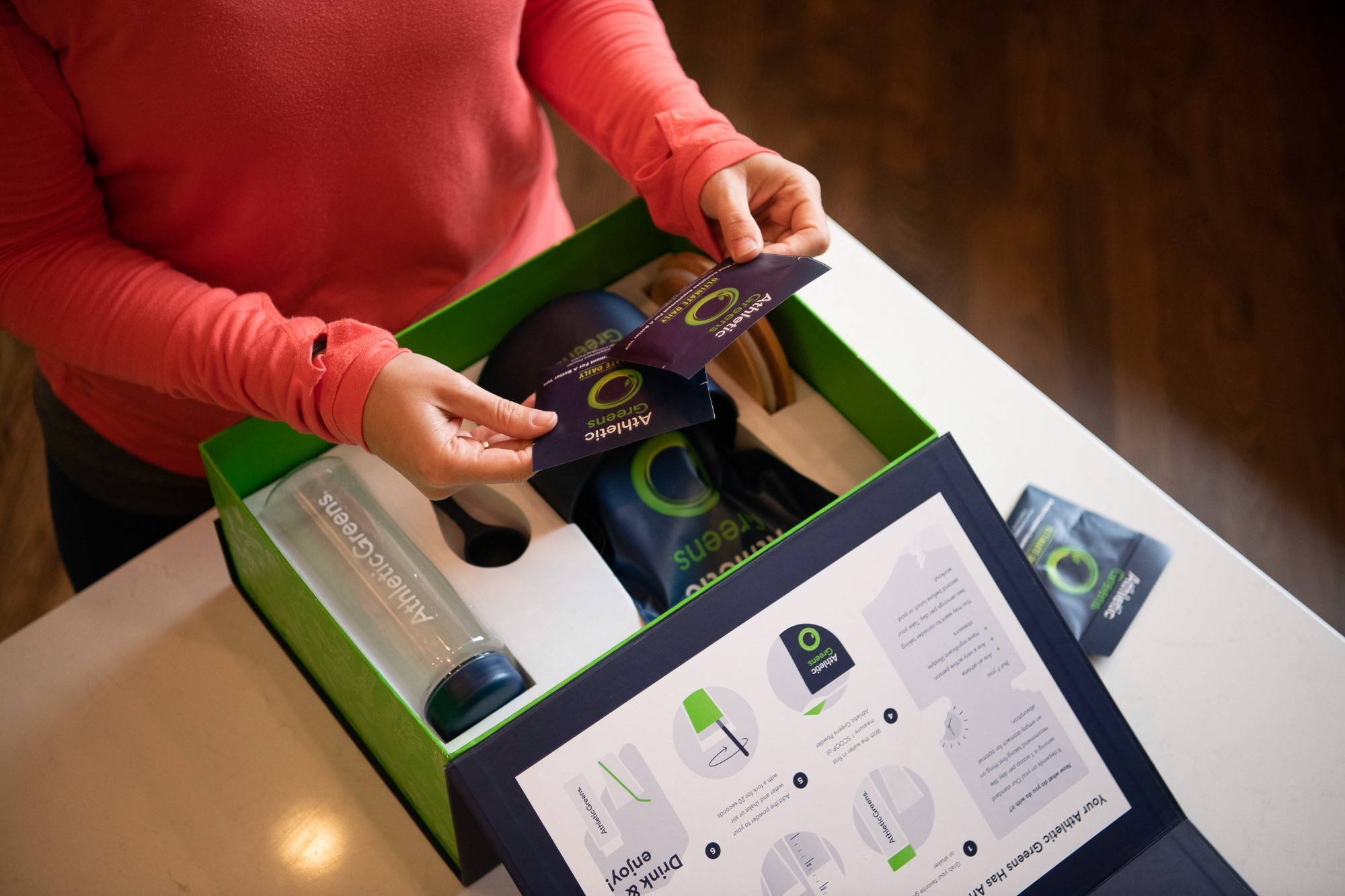 A person in a red sweater unpacks a green box while a tablet displays instructions nearby.
