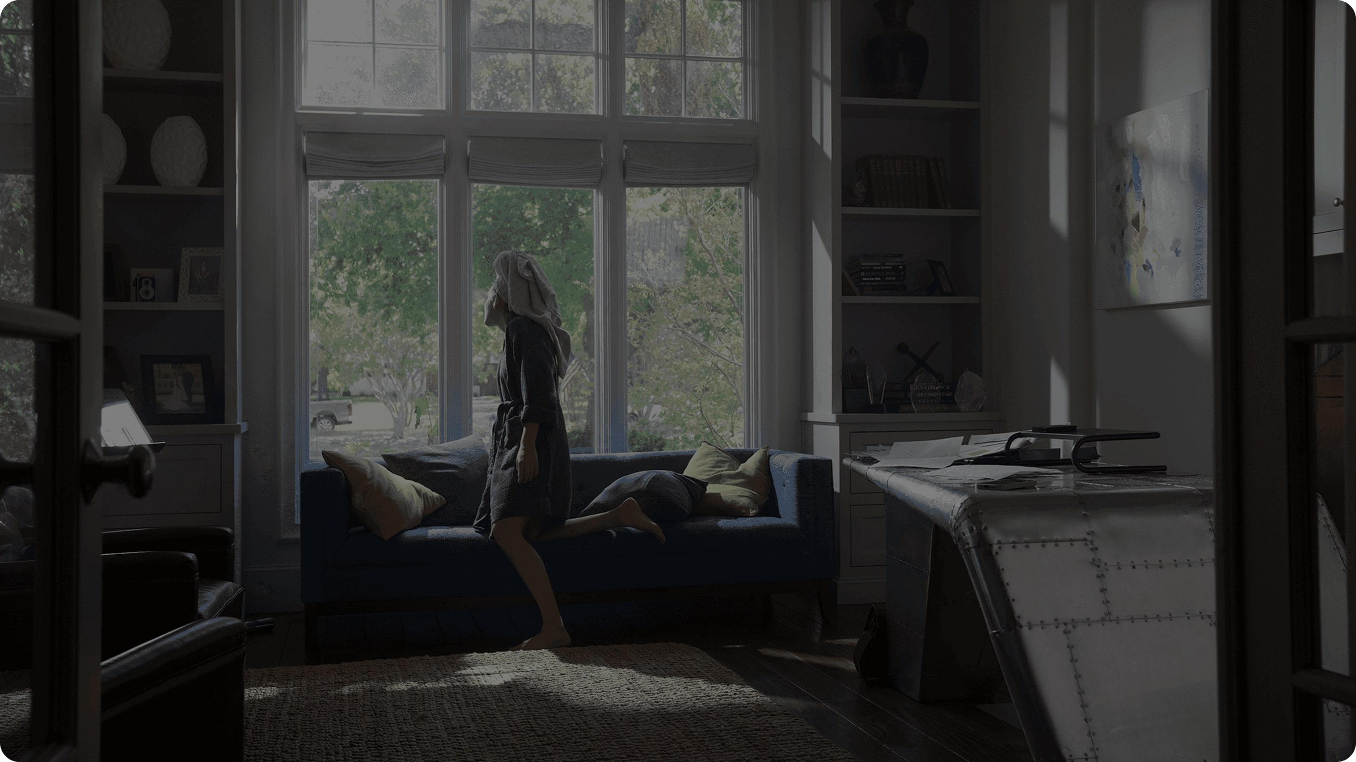 A person sits on the edge of a deep blue sofa in a well-lit living room, characterized by a large window with sheer curtains that let in natural light, surrounded by bookshelves and featuring a modern desk with metallic accents.