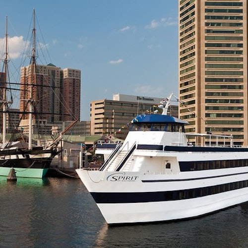 A large white yacht named "Spirit" docked in a harbor, with tall ships and city buildings in the background.