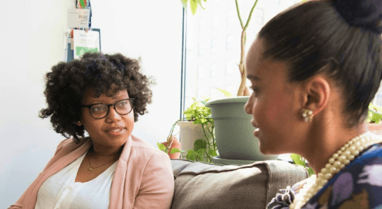 Two women sitting on a couch in a bright room, engaged in conversation.