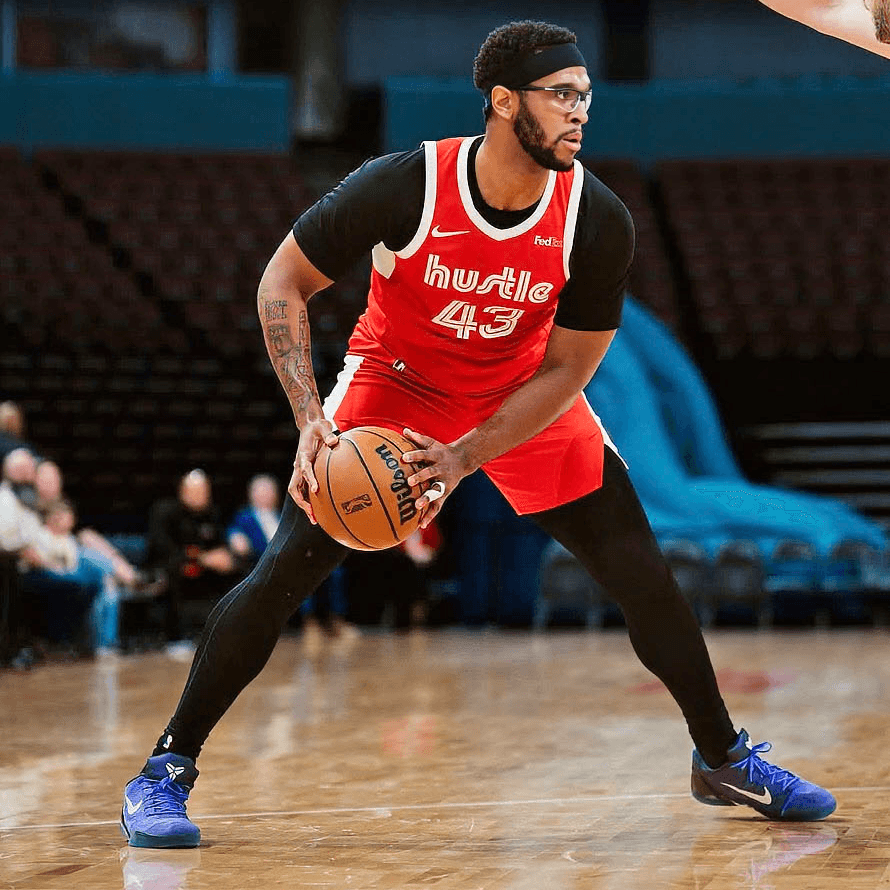 Eric Dixon in a red hustle jersey, wearing black leggings and blue shoes, dribbles the ball on a court while being guarded by an opponent, with empty seats visible in the background.