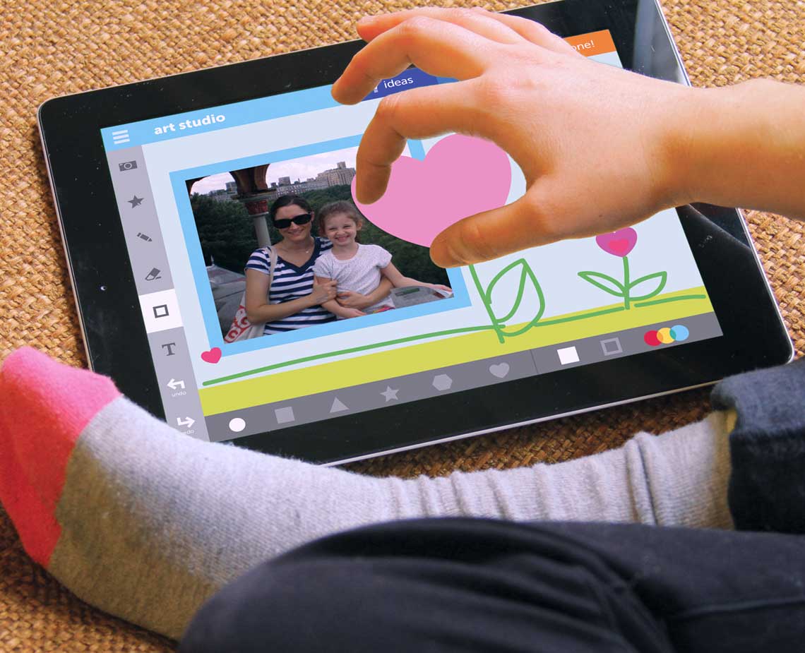 A child's hand holds a pink heart shape above a tablet displaying a photo of a woman and a child. The tablet screen shows a light blue border and icons for navigation, including a heart symbol and flower icons. The child is wearing gray socks with pink and gray stripes. The tablet rests on a textured brown surface.
