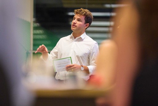 Smiling young woman with long hair standing against a dark green background, holding a finger to her chin.