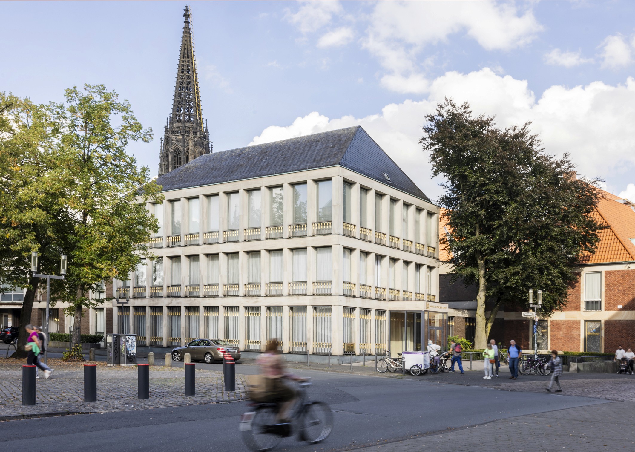 Modernes öffentliches Gebäude mit klarer Fassadenstruktur an einer belebten Straßenecke, umgeben von Bäumen, Radfahrenden und Passanten.
