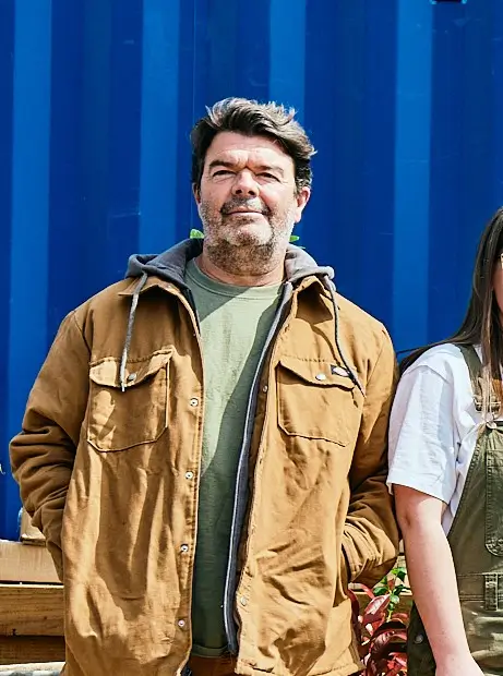 A person smiling, wearing a brown jacket, stands alongside another person against a blue background.