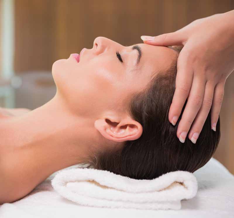 A woman lying down with a towel wrapped around her head, her face glowing with facial oil. She has a peaceful expression, enjoying a skincare treatment.