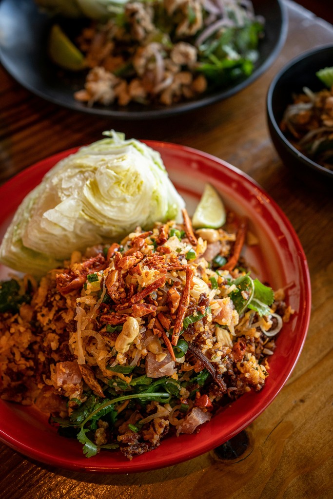 This vibrant image showcases a delicious plate of Thai larb, featuring crispy fried rice mixed with fresh herbs, vegetables, and topped with crunchy peanuts. The dish is served with a side of crisp lettuce and a lime wedge on a red-rimmed plate, perfect for wrapping the savory mix. The rich colors and textures highlight the dish's freshness and flavor, making it a mouthwatering option for those craving authentic Thai cuisine.