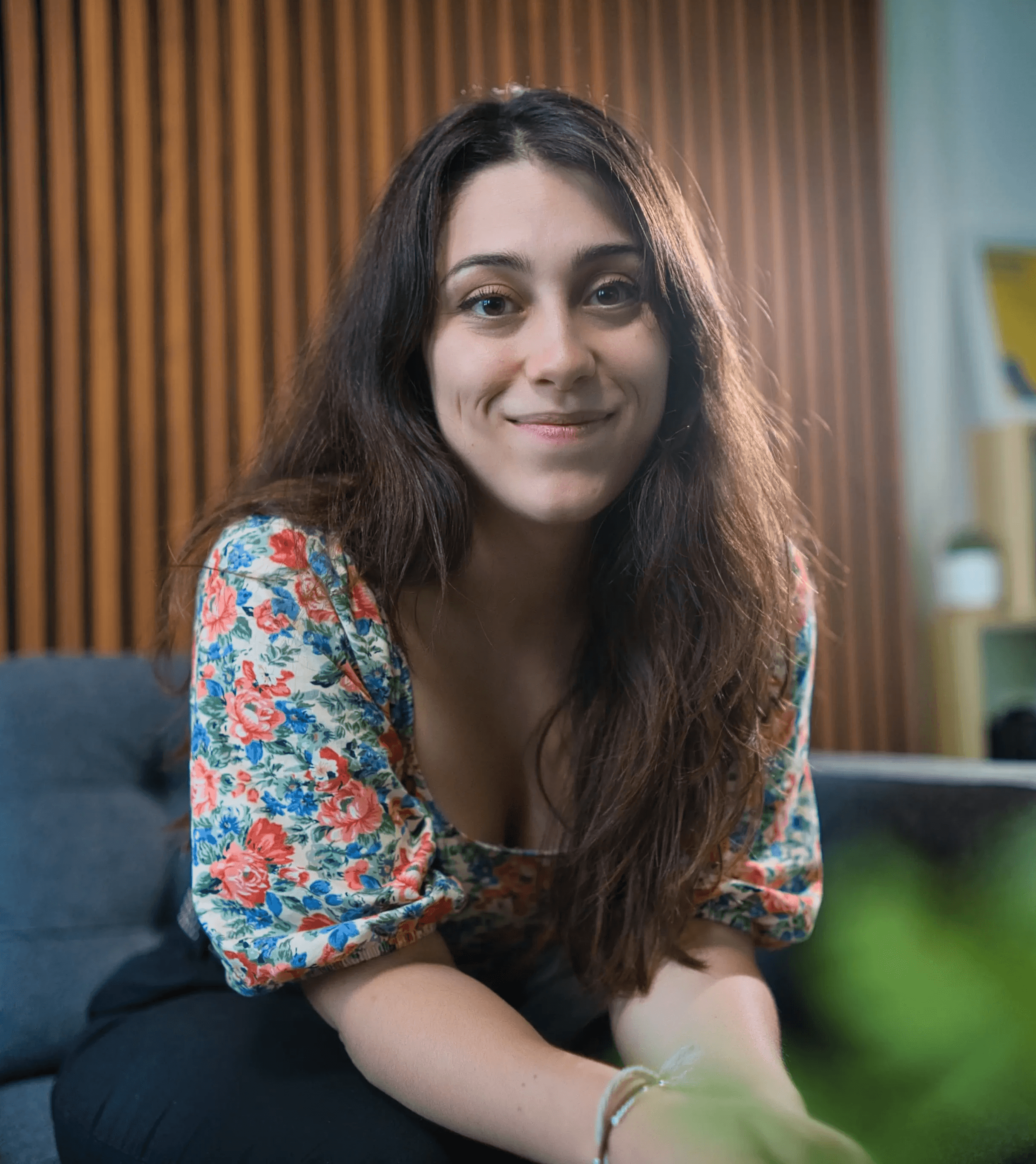 A woman with long brown hair and a floral patterned blouse sits on a gray sofa, framed by a wooden accent wall and soft, indoor lighting.