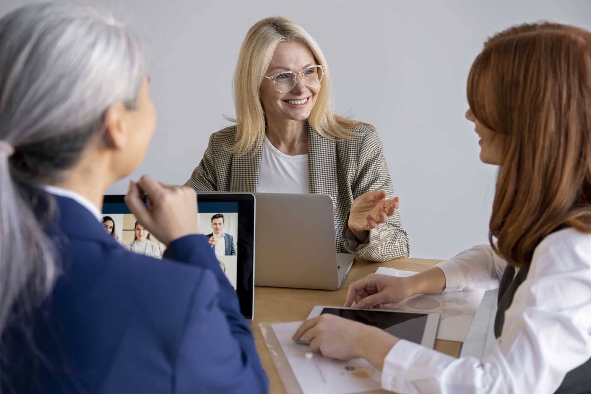 Three women in a collaborative meeting, with one blonde woman smiling and gesturing while discussing ideas, and a tablet displaying a video conference