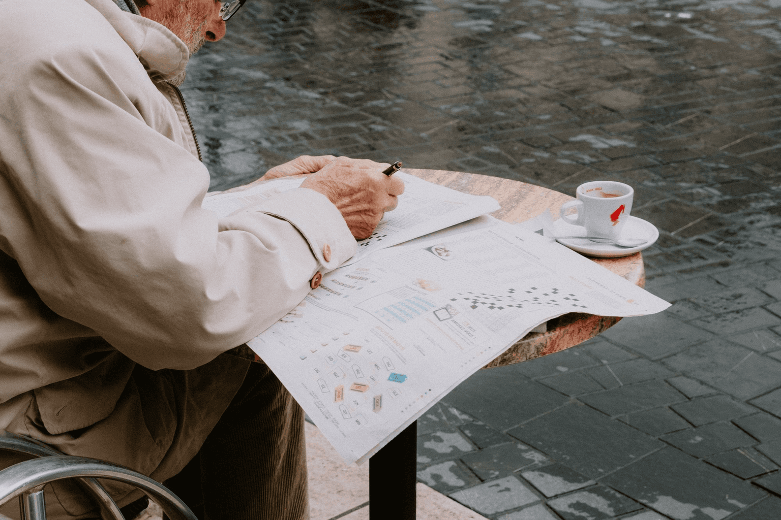 An older man filling in the newspaper puzzle page at a cafe table, an espresso beside him.