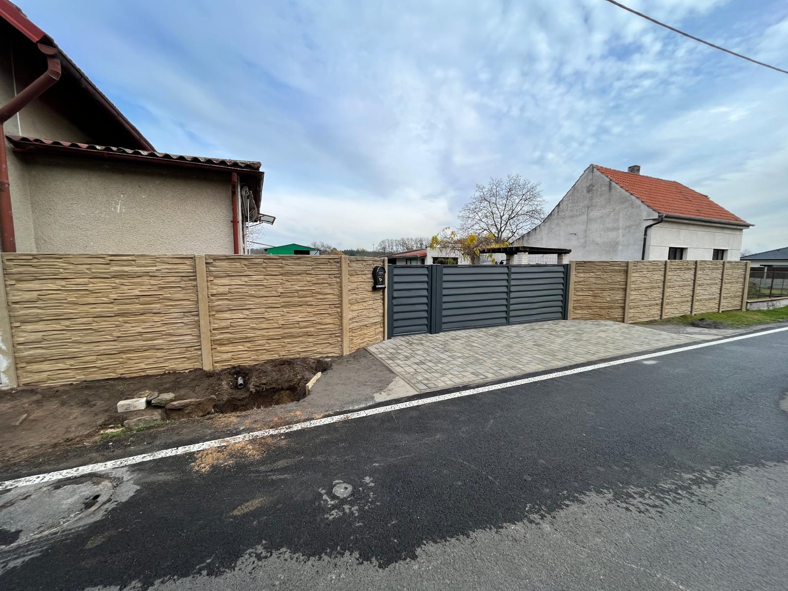 Předzahrádka s betonovým oplocením a kovovou bránou. Vedle domu a silnice. 