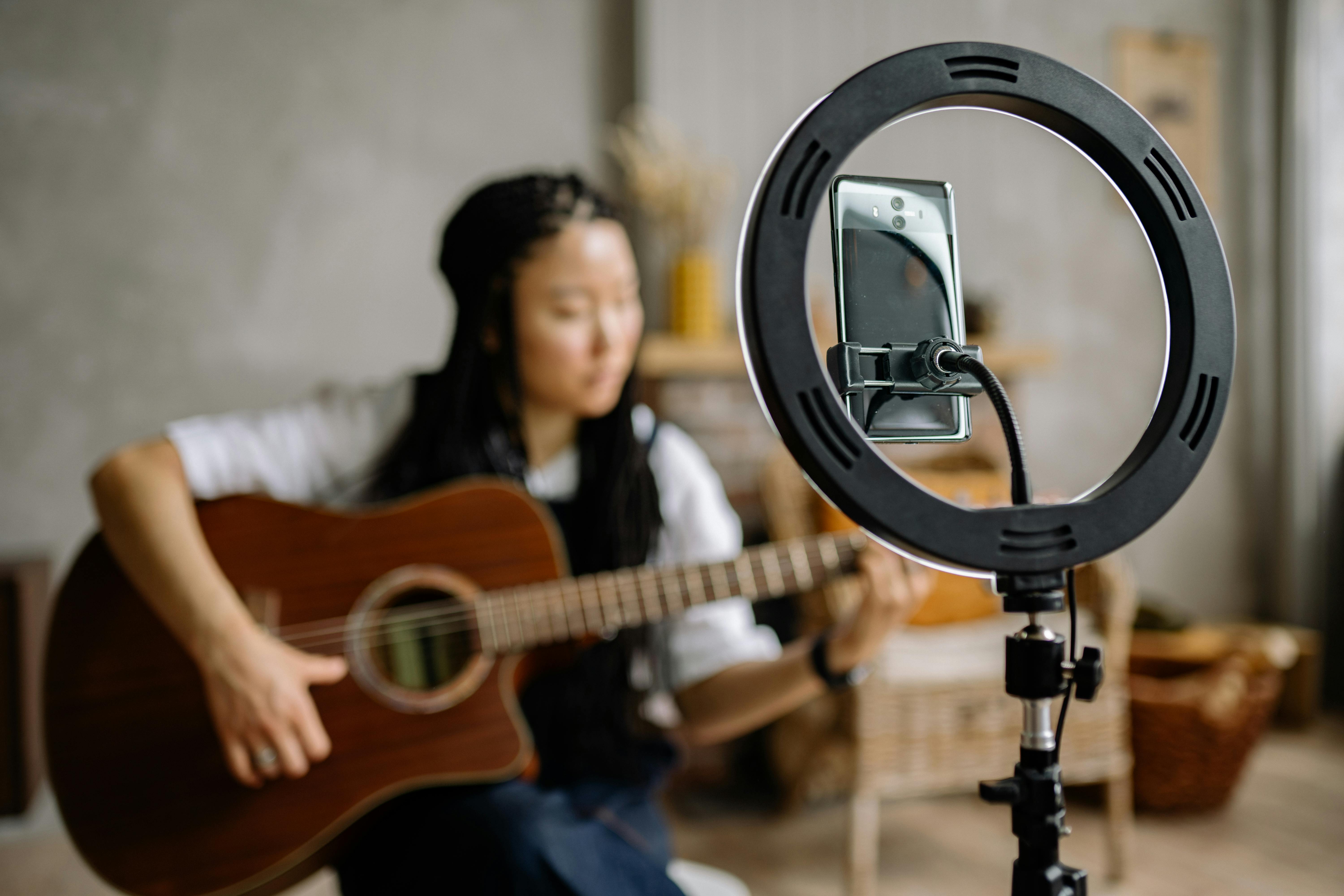 A phone on a tripod with a ring light frames a blurred person playing an acoustic guitar in a cozy indoor setting. The mood is creative and intimate.