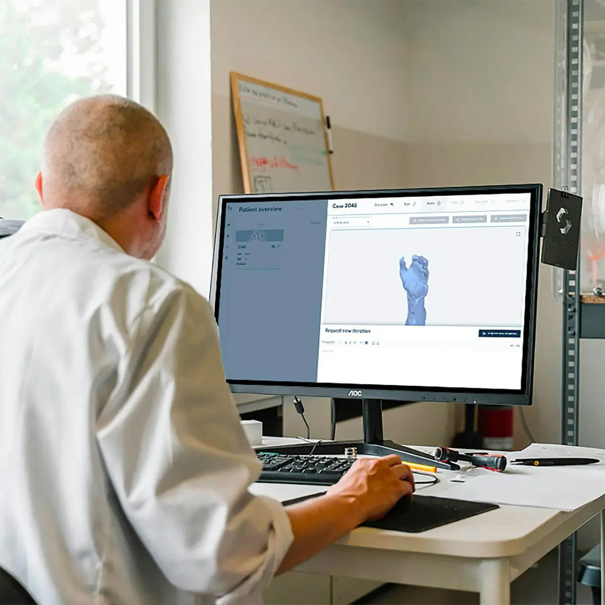 A man in a white lab coat working at a computer, viewing a 3D model on the screen in a laboratory or office setting.