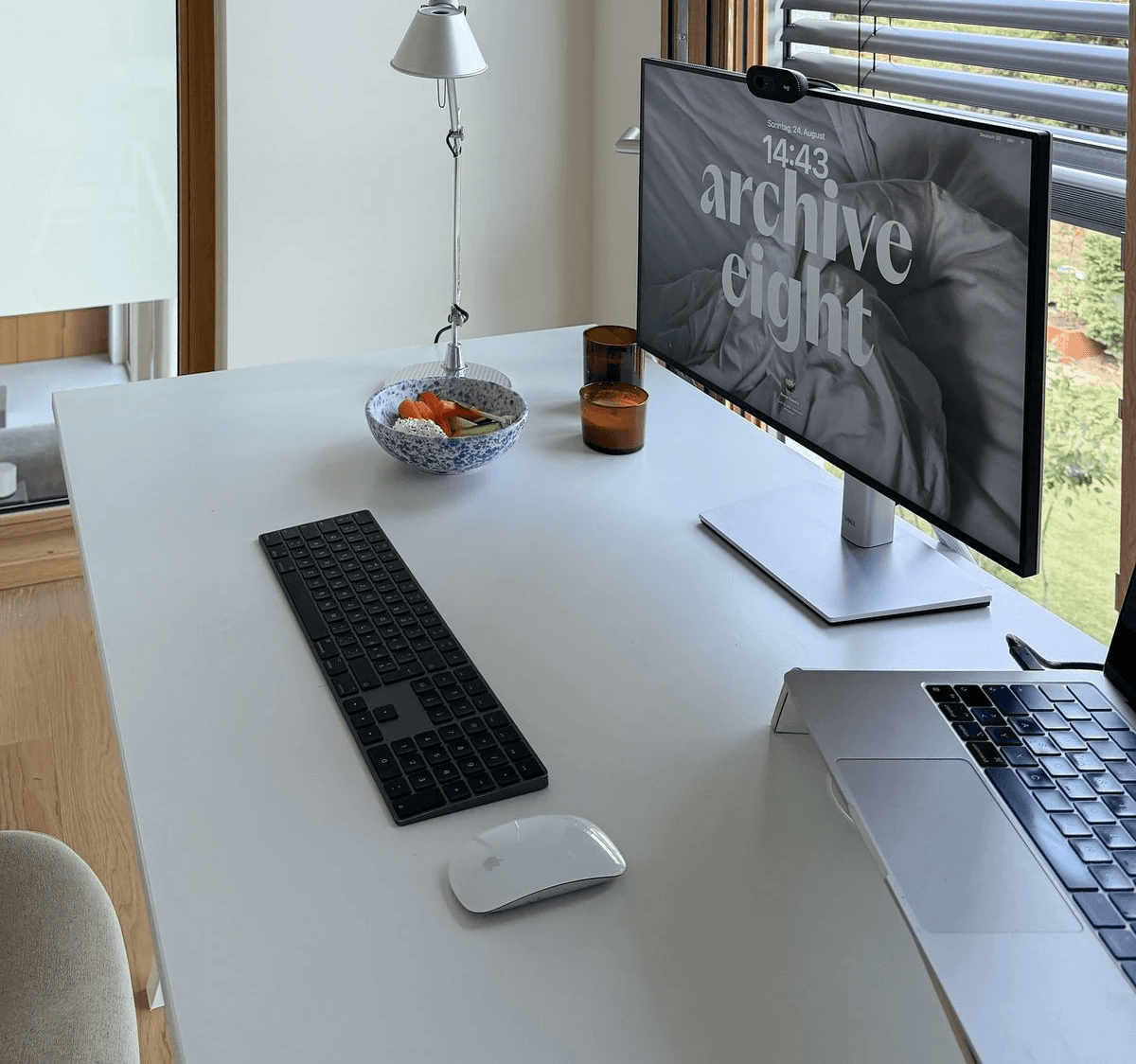 Modern minimalist desk setup with a computer monitor, microphone arm, camera, and indoor plants, featuring a pegboard with accessories on the wall and a Bauhaus-style poster leaning against the wall.