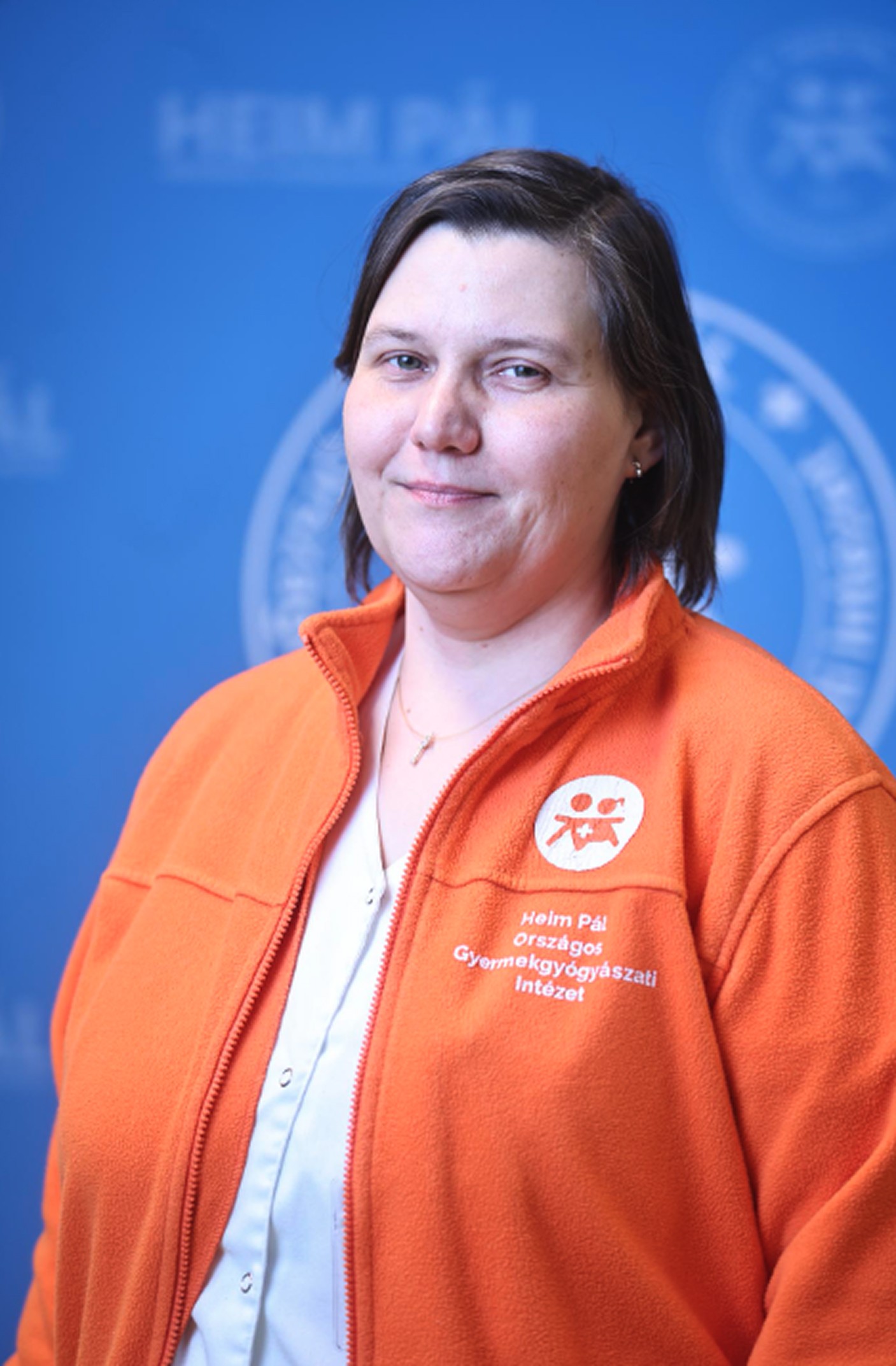 Hosszú barna hajú nő mosolyogva áll vörös ruhában egy kék, hivatalos fotófal előtt.