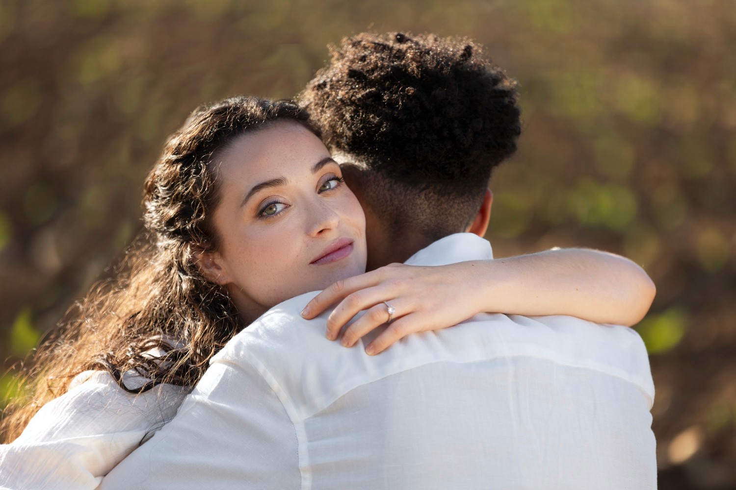 Woman hugging a man outdoors, looking at the camera. She wears an engagement ring. Romantic mood, blurred background.