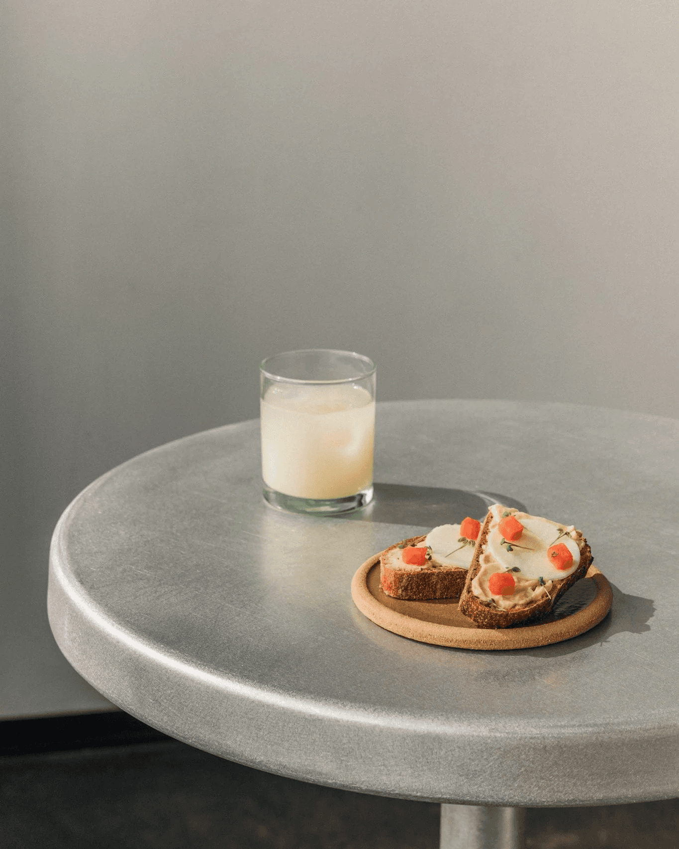 A round metal table holds a glass of milk and a slice of buttered bread topped with cherry tomatoes, both casting soft shadows.