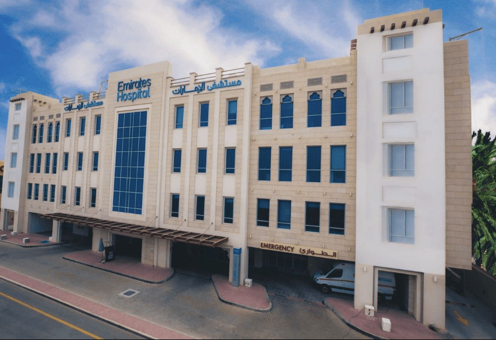 A modern hospital building in Dubai with a beige facade, large windows, and an emergency entrance at street level.