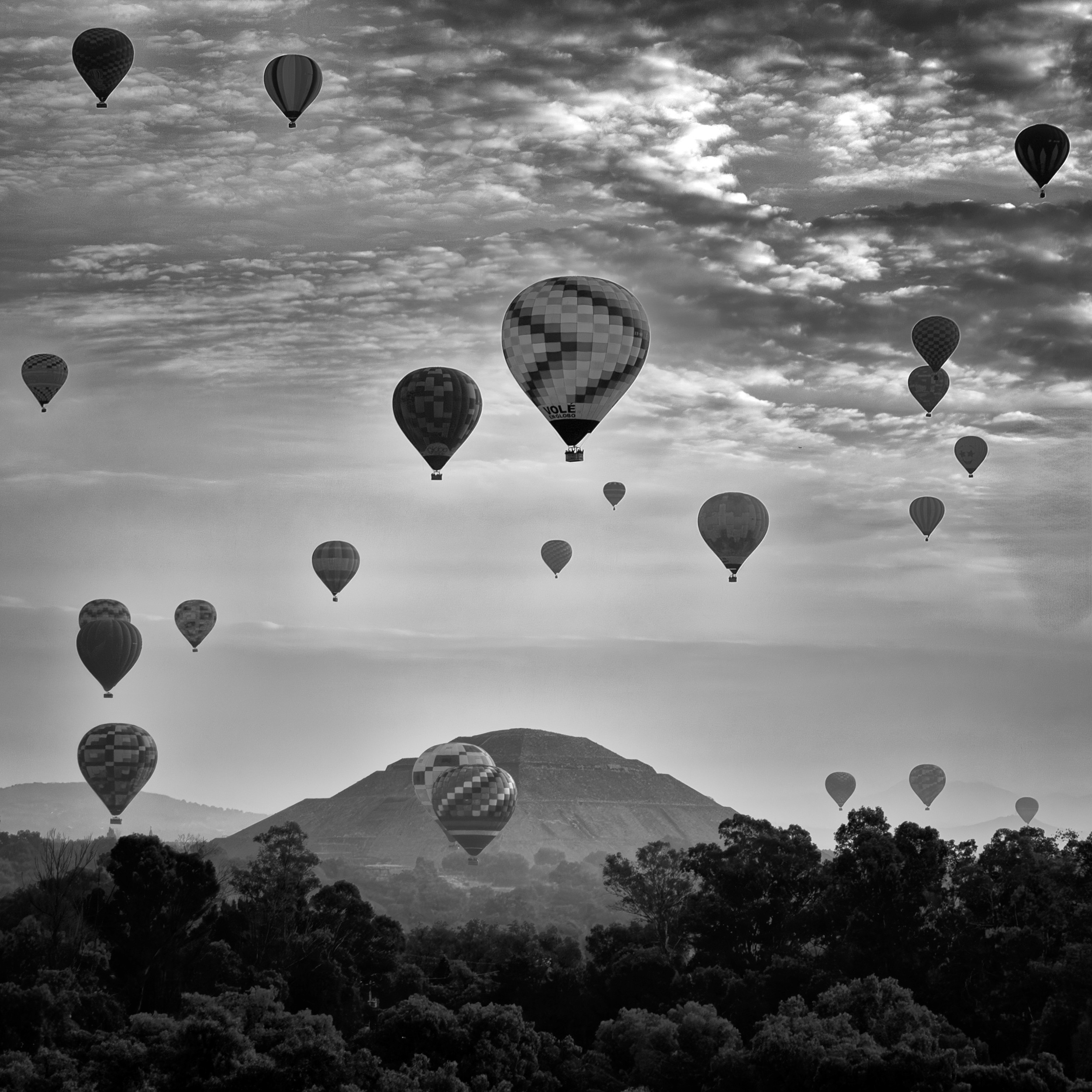 Black and white photo of numerous hot air balloons floating over the Pyramid of the Sun in Teotihuacán, Mexico, under a dramatic morning sky.